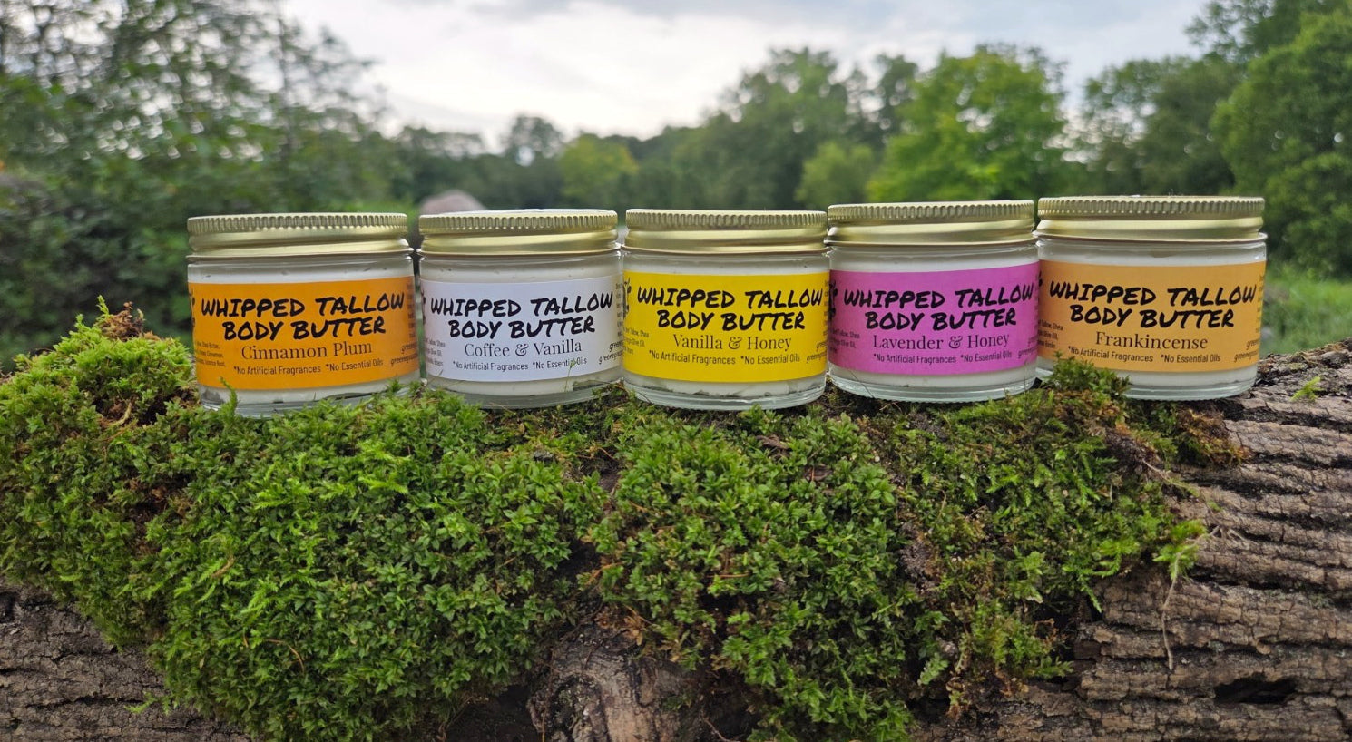 Five Greene Grass Tallow whipped moisturizer blends in glass jars on a mossy log.